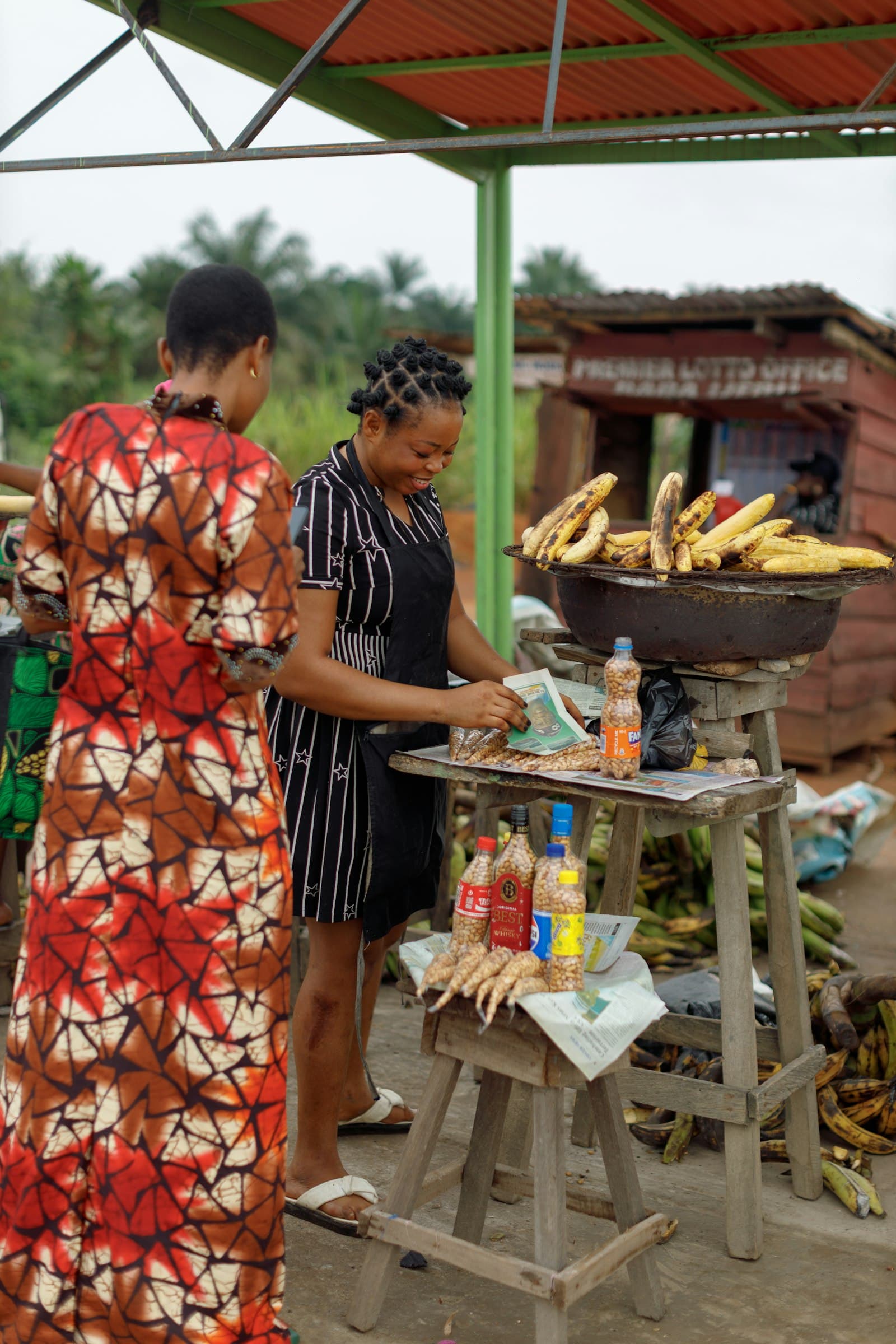 Two women at a Nigerian roadside stall selling plantain and snacks.