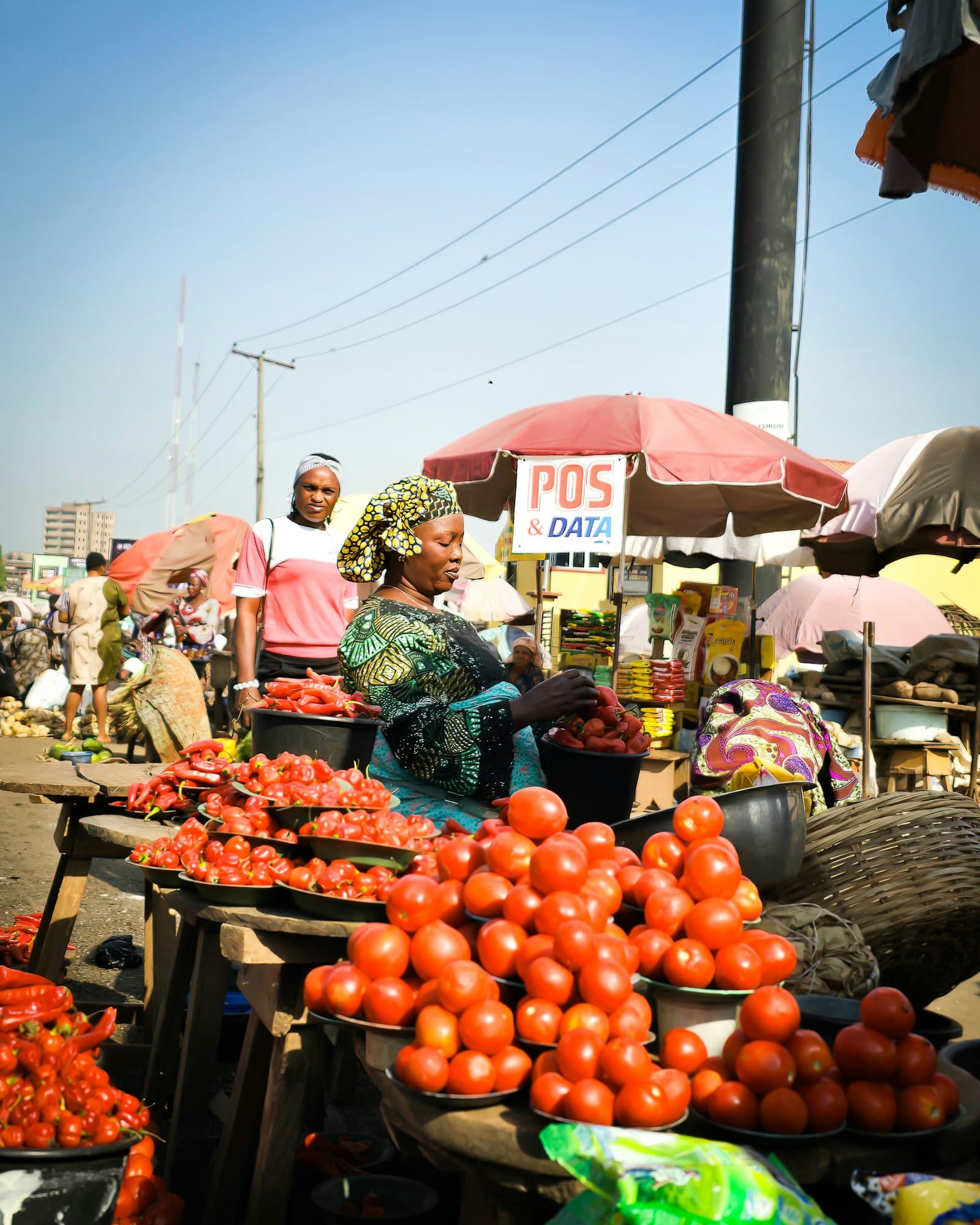 A Nigerian market trader at her stall of tomatoes and peppers, with a POS & DATA sign in the background.